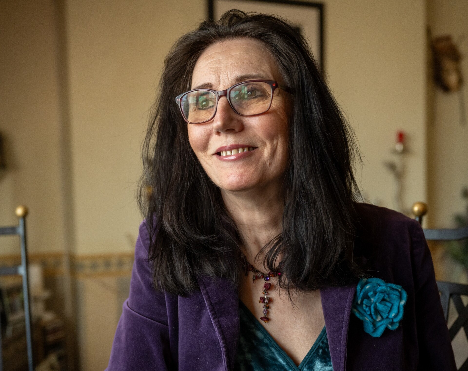 A portrait of Samantha Nelson wearing glasses, a purple velvet jacket with a blue fabric rose brooch, and a beaded necklace. 